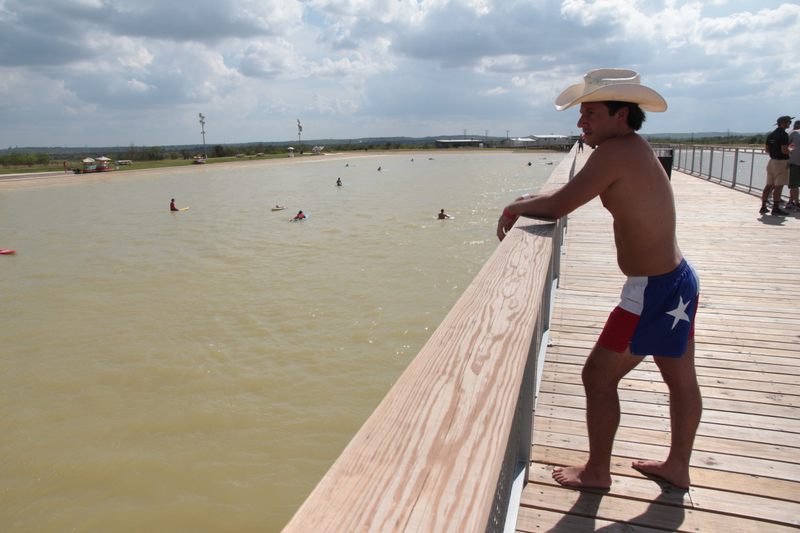 A man in Lonestar swim trunks looks out at surfers waiting for the next artificial wave at NLand Surf Park's grand opening in Austin, Texas.
